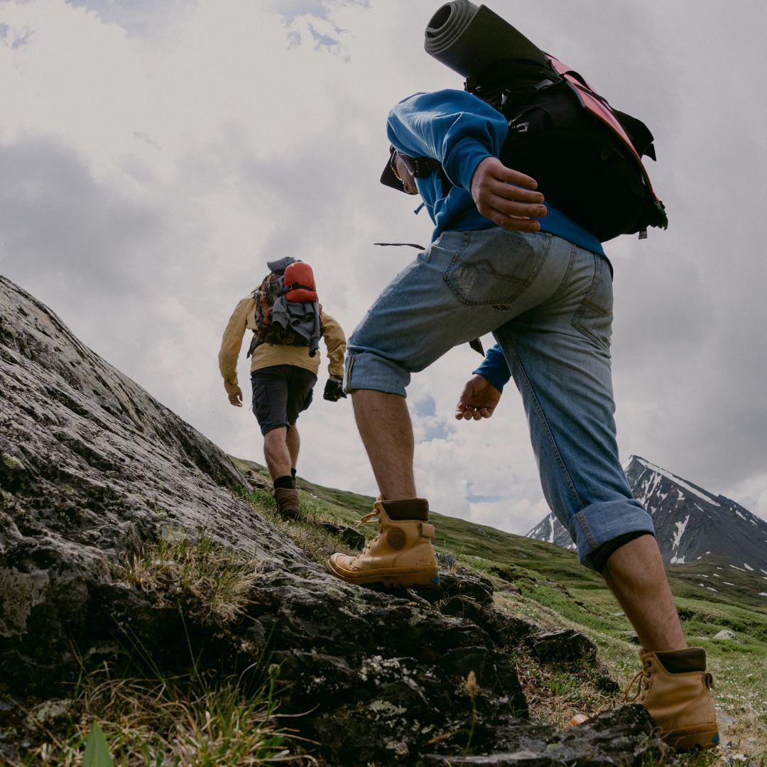 Two hikers ascend a rocky mountain slope under a cloudy sky. The foreground hiker wears a blue hoodie, light blue jeans, tan hiking boots, and carries a black backpack with a rolled sleeping pad.