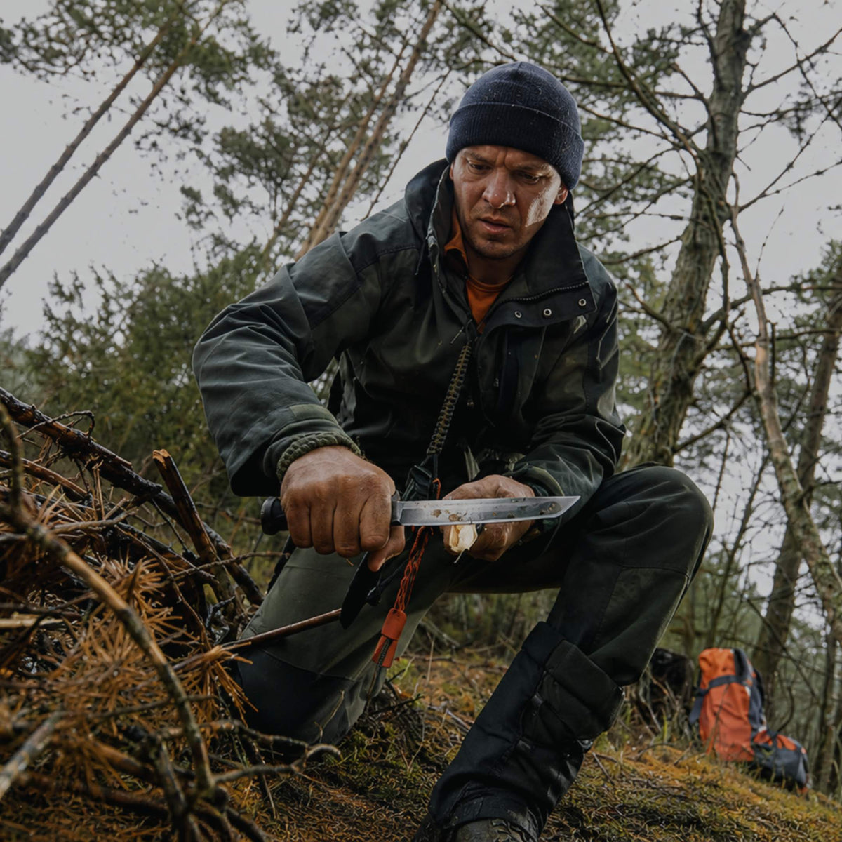 A man in outdoor gear, including a dark beanie, black jacket, and pants, is crouching in a forest while holding a knife. An orange backpack is visible in the background among trees.