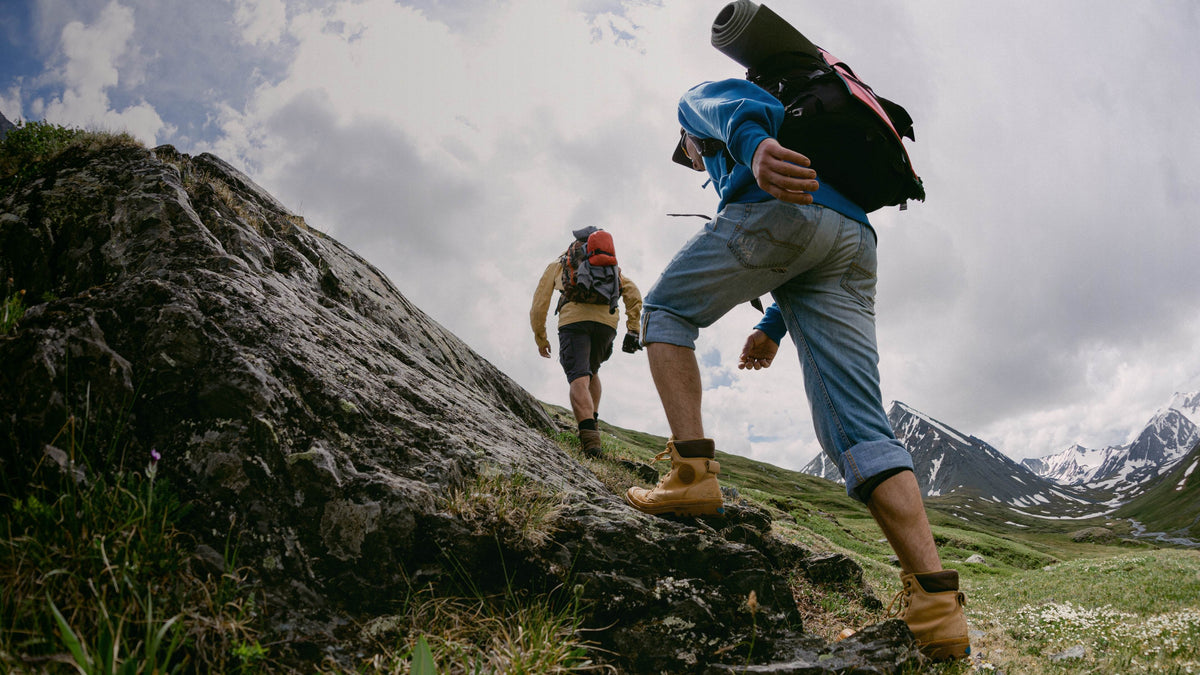 Two hikers ascend a steep rocky mountain slope under a cloudy sky, with snow-capped peaks and green alpine meadow in the background. The foreground hiker wears a blue jacket, rolled denim shorts, tan hiking boots, and a black backpack with a rolled sleeping bag, while the second hiker wears a beige top, dark shorts, and a red backpack.