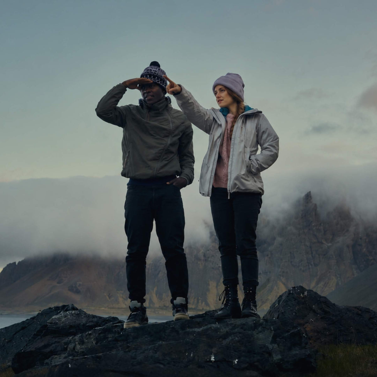 Two people standing on a rocky mountain peak with misty mountains and water in the background, wearing outdoor jackets and knit beanies.